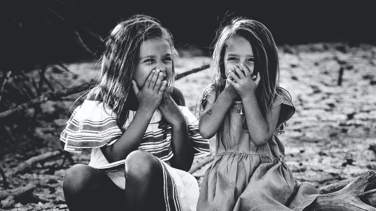 Bild vergrößern: zwei kleine Mädchen sitzen lachend nebeneinander und halten sich die Hände vor Ihr Gesicht. Bild in schwarz weiß.