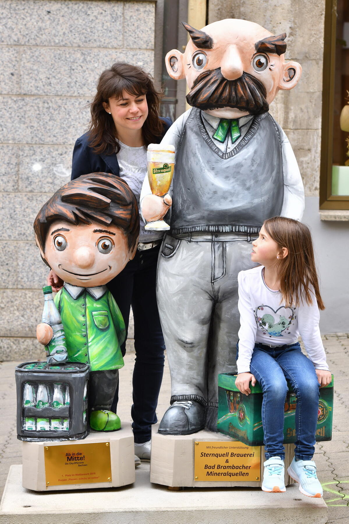 Bild vergrößern: Eine Frau steht und ein Mädchen sitzt bei den Figuren von Vater und Sohn, welche Getränke von der Sternquellbrauerei und den Bad Brambacher Mineralquellen halten.